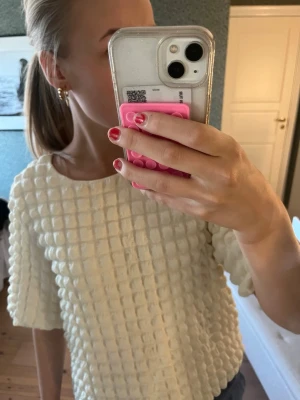 Vit topp med bubbligt strukturmönster - Säljer en vit topp med unikt bubbligt strukturmönster och rund halsringning. Toppen har halvlånga ärmar och är tillverkad i ett mjukt, texturerat material som ger en cool och trendig look. Perfekt för dig som vill sticka ut lite extra.