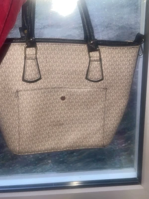 Beige Michael Kors handväska - Stilren beige handväska från Michael Kors med MK-logga över hela väskan. Väskan har svarta handtag och detaljer samt en stor ytterficka framtill. Insidan är fodrad i rosa tyg och har en innerficka med dragkedja. Perfekt för dig som gillar klassisk design och mycket plats. Har haft den i ett år men bara använt den 2 gånger.