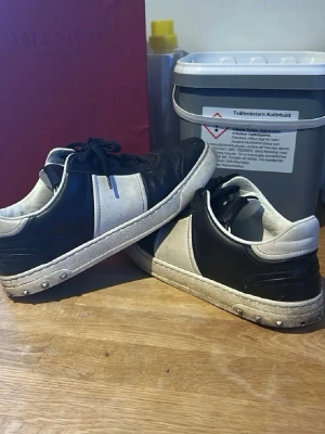 Svarta och vita sneakers från Valentino - Snygga sneakers från Valentino i svart och vitt läder med coola nitar längs sulan. Skorna har svarta skosnören och en vit panel på sidan. Yttersulan har ett unikt mönster och märkning undertill. Perfekta för dig som gillar streetstyle och designer. Passar 38,5