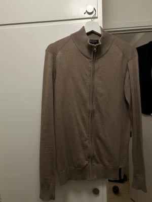 Beige ziptröja från Indicode Jeans - Snygg beige ziptröja från Indicode Jeans med hög krage och hel dragkedja framtill. Tröjan har ribbade muddar vid ärmslut och nederkant samt en diskret logolapp nedtill. Perfekt för lager på lager och enkel att matcha.