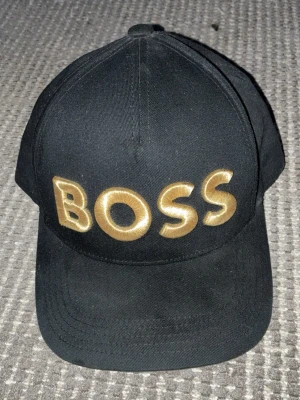 Svart keps med guldbrodyr BOSS - Snygg svart keps från BOSS med stor guldfärgad brodyr på framsidan där det står 'BOSS'. Klassisk böjd skärm och justerbar baksida. Tillverkad i slitstarkt tyg som ger en clean och modern look kom privat för frågor