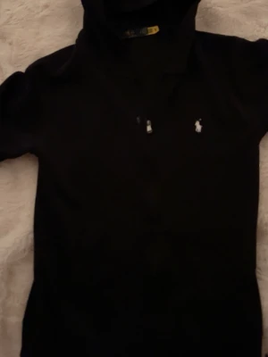Svart hoodie från Polo Ralph Lauren - Snygg svart hoodie från Polo Ralph Lauren med klassisk vit broderad logga på bröstet. Hoodien har huva och är tillverkad i mjukt material som känns skönt mot huden. Perfekt för dig som gillar stilrena och tidlösa plagg.