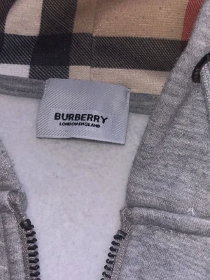 Grå zip hoodie från Burberry - Snygg grå hoodie från Burberry med dragkedja och klassiskt rutigt foder i huvan. Tröjan har fickor framtill, justerbar huva med snören och diskret Burberry-logga på bröstet. Perfekt för en chill och stilren look.