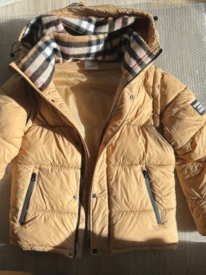 Beige pufferjacka med rutigt foder - Stilren beige pufferjacka från Burberry med klassiskt rutigt foder i huvan. Jackan har två dragkedjefickor framtill, huva med dragsko och patch på ärmen. Perfekt för kalla vinterdagar och ger en lyxig känsla till din outfit.