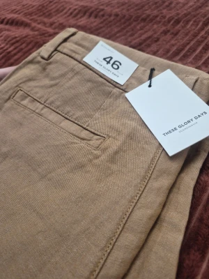 These Glory Days T Fairfox konjak färg byxor, storlek 46 - Snygga beige chinos från These Glory Days med klassisk rak passform och diskreta fickor både fram och bak. Tillverkade i ett mjukt bomullstyg som känns bekvämt hela dagen. Perfekta för dig som gillar stilrena och tidlösa plagg.