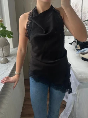 Svart halterneck topp med spets zara - Säljer en svart halterneck topp i satinliknande material med spetsdetaljer längs kanterna. Toppen är ärmlös och har en asymmetrisk nederkant som ger en cool och trendig look. Perfekt att styla med jeans eller kjol för en snygg outfit.