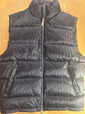 Svart dunväst från Dior - Snygg svart dunväst från Dior med diskret logomönster över hela västen och hög krage. Västen har dragkedja framtill och två fickor. Perfekt för att lägga till ett exklusivt lager till din outfit.