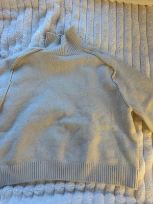 Beige stickad tröja med hög krage - Säljer en beige stickad tröja med hög ribbad krage och långa ärmar. Tröjan har bred ribbstickad mudd vid nederkant och ärmar, samt en avslappnad passform. Perfekt för kalla dagar och enkel att styla till olika outfits.