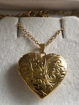 Guldfärgat hjärthalsband medaljong med mönster - Snyggt halsband i guldfärg med ett hjärtformat medaljonghänge. Hänget har ett detaljerat, blommigt mönster och kan öppnas för att lägga in en liten bild eller minne. Kedjan är tunn i rostfritt stål och elegant, perfekt för att ge din stil en personlig touch.