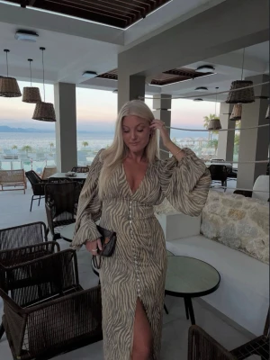 Adoore klänning  - Superfin klänning , 1 knappar längst ner har försvunnit men går lätt att sy på en ny knapp, annars fint skick. Adoore Postiano den långa modellen, zebra mönster som varit slutsåld ett bra tag.  Positano maxi dress in a brown zebra pattern. The dress has buttons and slits at the front, bold long sleeves, and a V-necked collar. It is lined, has a hidden zipper at the back, and is full-length. 