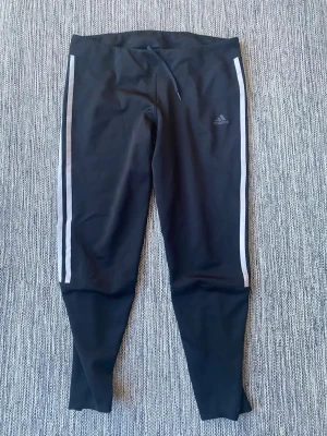Svarta Adidas leggings med ränder - Adidas Techfit träningstights för dam i storlek L. Långa tights i svart färg med elastisk midja. Tillverkade i ett stretchigt funktionsmaterial som ger stöd vid träning. Modellen har paneler i mesh och tryckt Adidas-logga på benet. Passar för olika typer av träning och löpning.  Använda och lite noppriga precis på sömnen i övrigt i bra begagnat skick   Mått Över midjan: ca 38 cm Längd från grenen och ner: ca 73 cm  Kommer från ett djur och rökfritt hem  