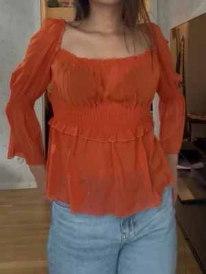 Orange transparent blus med puffärm - Trendig orange blus i transparent material med smockad midja och volangdetaljer. Blusen har fyrkantig ringning och puffiga långärmar som ger en romantisk vibe. Perfekt att styla med jeans för en cool look.