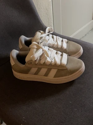 Beige Adidas sneakers med vita detaljer - Snygga beige sneakers från Adidas med klassiska vita ränder på sidan och breda vita skosnören. Ovandelen är i mockaliknande material och sulan är vit. Perfekta för dig som gillar stilrena och tidlösa sneakers med en sportig vibe.