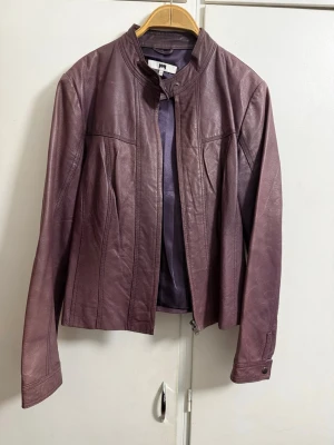 Lila skinnjacka från Joy - Snygg lila skinnjacka från Joy med rak passform och ståkrage. Jackan har dragkedja framtill och dekorativa sömmar som ger en cool look. Perfekt för dig som vill sticka ut med en unik färg på din skinnjacka.