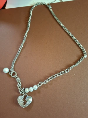 Silverfärgat halsband med pärlor och hjärta - Snyggt silverfärgat halsband med grov kedja, dekorerat med vita pärlor och ett glittrande hjärthänge med blixtformad spricka. Perfekt accessoar för att lyfta din outfit med en edgy touch.