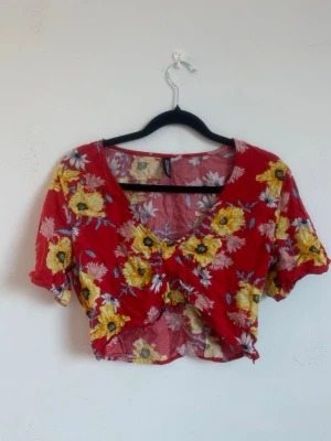 Röd blommig croppad topp H&M - Snygg croppad topp från H&M i rött med blommigt mönster i gult, vitt och blått. Toppen har korta ärmar, v-ringning och detalj framtill. Perfekt för sommarens alla tillfällen och ger en färgglad vibe. Liten i storlek!
