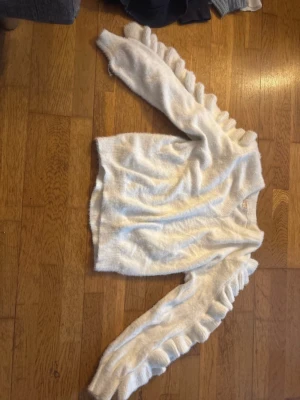 Vit fluffig stickad tröja med volanger - Supersöt vit stickad tröja med volangdetaljer längs ärmarna. Tröjan är fluffig och mjuk, tillverkad i 50% polyamid och 50% akryl. Perfekt för dig som vill ha något extra till din outfit.