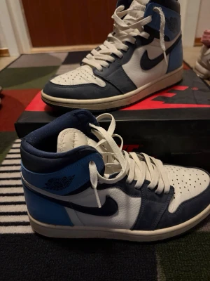 Nike Air Jordan 1 obsidian - Säljer ett par Nike Air Jordan 1 High sneakers i blått, vitt och marinblått. Skorna har klassisk hög siluett, vit tåbox och sidor, mörkblå detaljer och ljusblå häl. Tillverkade i läder med ikoniska Swoosh-loggan på sidan och Air Jordan Wings på ankeln.
