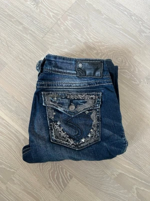 Mörkblå bootcut jeans med bling - Säljer ett par mörkblå bootcut jeans från Silver med  snygga detaljer på bakfickan. Fickan har silverfärgade nitar, broderier och bling som ger extra attityd. Klassisk femficksmodell i stretchigt denimtyg.