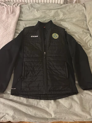 Svart jacka från CCM med klubbmärke - Svart jacka från CCM med klubbmärke på bröstet och dragkedja framtill. Ångermanland ishockeyförbund jacka ifrån tv-pucken. Jackan är använd men i topp skick utan några skador eller fläckar. 