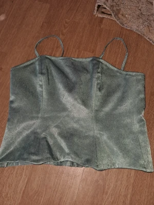 Grön satin linnetopp med smala band - Snygg grön linnetopp i satin med smala axelband och rak passform. Perfekt att styla med jeans eller kjol för en trendig look. Materialet har en lätt glansig finish som ger en lyxig känsla.