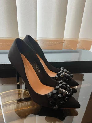 Svarta pumps med stora pärlor - Snygga svarta pumps från BIGTREE med spetsig tå och höga klackar. Skorna har en dekorativ detalj framtill med stora svarta pärlor och en silvrig accent. Materialet ser ut att vara satinliknande tyg som ger en elegant finish.