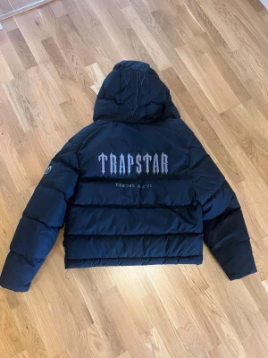 Svart pufferjacka från Trapstar - Säljer en svart Trapstar jacka med logga på ryggen och mindre tryck på huvan och framsidan. Storlek M