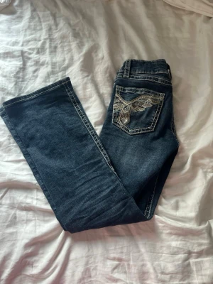 Mörkblå bootcut jeans med brodyr - Säljer ett par mörkblå bootcut jeans med vita kontrastsömmar och snygg broderad bakficka med detaljer i silver. Jeansen har låg midja och klassisk femficksdesign. Perfekta för dig som gillar en avslappnad men ändå trendig look. Storlek 146