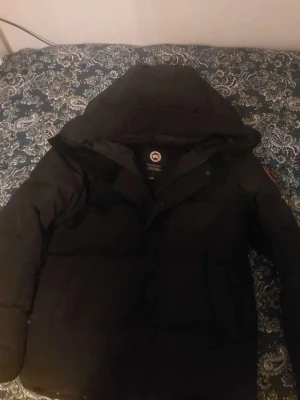 Svart dunjacka från Canada Goose - Säljer en svart dunjacka från Canada Goose med huva och klassisk logotyp på ärmen. Jackan är tjockt vadderad och har en stilren design som håller dig varm under kalla vinterdagar. Perfekt för dig som vill ha både funktion och stil. (FAST PRIS)