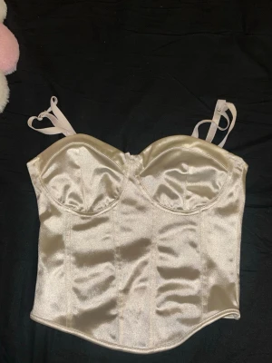 Champagnefärgad satin korsettopp - Snygg vit färgad  korsettopp i satin med reglerbara axelband och markerade kupor. Toppen har en glansig finish och ger en elegant siluett. Perfekt att styla med jeans eller kjol för en trendig look.