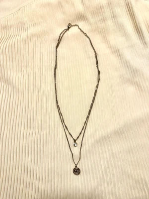 Dubbelhalsband med mynt och sten - Guldigt halsband som ser ut som två stycken i ett, det långa med litet mynt och det kortare med sten. Använt 1-3 ggr så säljes utan tecken på användning.