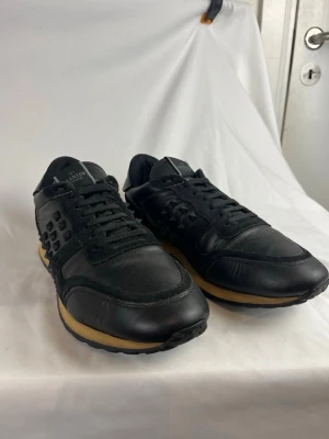 Valentino Rockrunners  - Snygga svarta sneakers från Valentino med nitar på sidorna och bak. Skorna har en svart ovandel i läder och mocka, svarta skosnören och en beige sula med svarta detaljer. Perfekta för dig som gillar en edgy och exklusiv stil.