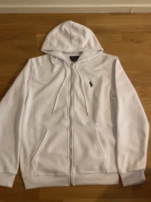 Vit hoodie från Polo Ralph Lauren - Snygg vit hoodie med dragkedja från Polo Ralph Lauren. Klassisk design med huva, dragsnören och kängurufickor framtill. Liten broderad logga på bröstet. Perfekt för en clean och stilren look.