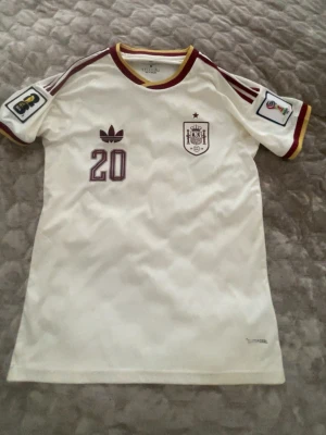 Vit Spanien matchtröja Adidas Pedri - vm 2026 - Säljer en vit Spanien matchtröja från Adidas med vinröda detaljer, nummer 20 och namnet Pedri på ryggen. Tröjan har officiella VM-märken på ärmarna, klubbmärke på bröstet och klassiska Adidas-ränder på axlarna. Perfekt för fotbollsfans! OBS har en liten fläck som knappt syns (visas på bild).