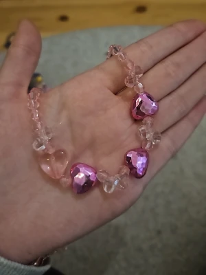 Rosa halsband med hjärtformade pärlor - Sött halsband med rosa och genomskinliga plastpärlor, dekorerat med tre större hjärtformade pärlor i metallicrosa och en i ljusrosa. Halsbandet har ett plastlås och pärlorna är trätt på elastisk tråd. Perfekt för dig som gillar gulliga accessoarer och hjärtan.