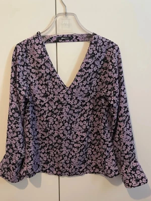 Lila blommig blus från Gina Tricot - Supersnygg blus från Gina Tricot i svart med lila blommönster. Djup V-ringning framtill och långa ärmar. Tunt och mjukt tyg som känns skönt mot huden. Perfekt att styla med jeans eller kjol för en trendig look.