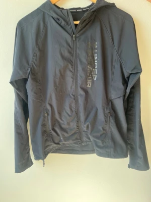 Svart vindjacka från Under Armour - Snygg svart vindjacka från Under Armour med huva och hel dragkedja framtill. Jackan har två fickor med dragkedja och ett stort Under Armour-tryck på bröstet. Tillverkad i lätt och slitstarkt syntetmaterial, perfekt för träning eller utomhusaktiviteter.