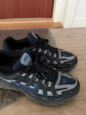Nike Air Max mörkblå/svarta sneakers - Nike Air Max sneakers i mörkblått och svart med grå detaljer. Skorna har mesh- och syntetpaneler, klassisk Nike Swoosh i blått på sidan och svarta snören. Låg modell med robust sula och sportig vibe. Perfekta för dig som gillar streetwear och retrostil.