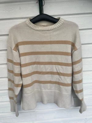 Beige randig stickad tröja Stories - Stickad tröja från Stories i beige med ljusbruna horisontella ränder. Tröjan har rund halsringning, långa ärmar och bred ribbstickad kant nedtill. Perfekt för dig som gillar en avslappnad men ändå stilren look. Den är väl använd och lite missfärgad men ej nåt man tänker på när man har på sig den