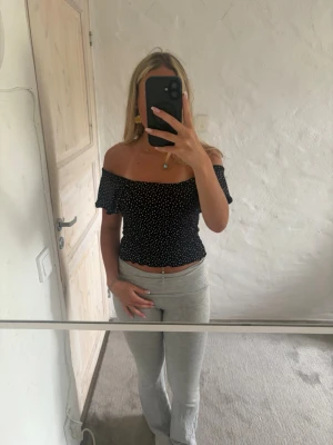 Svart offshoulder topp med prickar - Snygg svart offshoulder topp med vita prickar och volangiga ärmar. Croppad modell med stretchig passform och vågig kant nedtill. Perfekt för dig som vill ha en trendig och bekväm topp till sommaren.