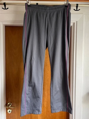 Grå capri track pants med rosa detaljer - Snygga grå track pants i capri längd. Mid till low rise midja, smala ben och elastisk midja. Byxorna har tunna rosa ränder längs sidorna som ger en sportig vibe. Perfekta för chill dagar eller när du vill ha det bekvämt. Tillverkade i mjukt bomullsmaterial.