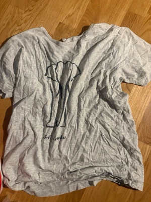 Grå t-shirt med elefantmotiv - Säljer en ljusgrå t-shirt med ett stilrent svart elefantmotiv och texten 'Sri Lanka' på ryggen. T-shirten har korta ärmar och är gjord i mjuk bomull, perfekt för en avslappnad look.