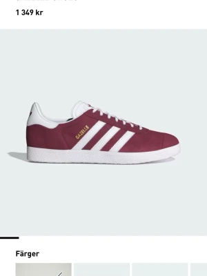 Adidas Gazelle vinröda sneakers - Säljer ett par klassiska Adidas Gazelle sneakers i vinrött mocka med vita detaljer och de ikoniska tre ränderna på sidan. Skorna har vit sula och snörning samt guldfärgad Gazelle-text på sidan. Perfekta för en stilren och sportig look.