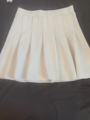 Vit plisserad kortkjol - Snygg vit kortkjol med plisserad design och bred linning. Kjolen har en klassisk och stilren look som passar perfekt till många olika outfits. Materialet känns lätt och bekvämt.