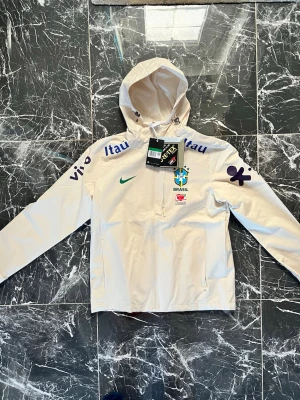 Vit Brasil GORE-TEX vindjacka Nike - Snygg vit vindjacka från Nike med Brasil-logga och flera sponsortryck. Jackan har huva med dragsko, hel dragkedja framtill och två sidofickor. Tillverkad i GORE-TEX-material som står emot regn och vind. Perfekt för dig som gillar sportig stil och vill sticka ut.