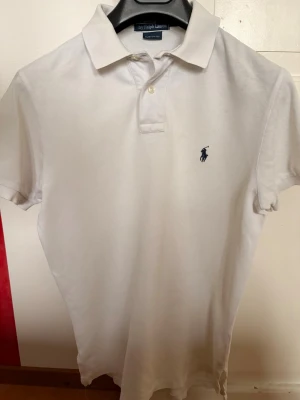 Vit pikétröja från Ralph Lauren - Klassisk vit pikétröja från Ralph Lauren med korta ärmar och blå broderad logga på bröstet. Tröjan har krage och knappar vid halsen, tillverkad i mjuk bomull som känns skön mot huden. Perfekt för en clean och stilren look.
