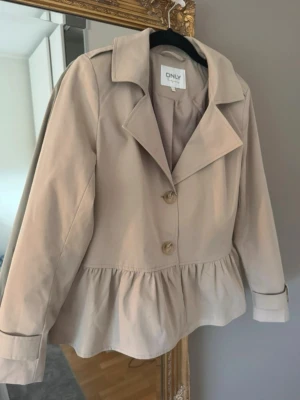 Beige trenchcoat från ONLY - Stilren beige kavajjacka från ONLY med volang nedtill och stora knappar framtill. Jackan har klassisk krage, lång ärm och markerad axel. Perfekt för dig som vill ha en snygg kappa till höst och vår. 