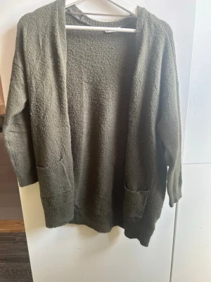 Olivgrön mysig kofta med fickor - En oversized olivgrön kofta med två stora fickor framtill. Koftan är stickad i ett mjukt, fluffigt material och har långa ärmar samt ribbade muddar vid ärmslut och nederkant. Perfekt för lager på lager och chill dagar.