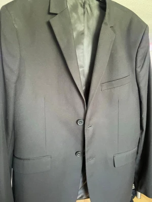 Kavaj från H&M - Stilren svart kavaj från H&M i slim fit-modell. Kavajen har två knappar framtill, klassisk krage och tre fickor. Fodret är slätt och glansigt för en snygg finish. Perfekt för dig som gillar en modern och enkel look.
