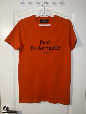 Orange Peak Performance t-shirt - Snygg orange t-shirt från Peak Performance med svart tryck framtill. Klassisk rund halsringning och korta ärmar. Perfekt för en avslappnad och sportig stil. Finns att hämta i Södertälje.